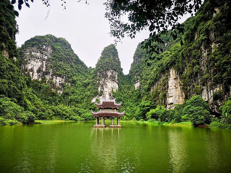 A small temple sits on the edge of a small body of water, surrounded by tall, tree-covered limestone mountains. In Trang An, Ninh Binh, Vietnam